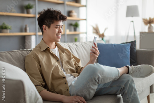Smiling asian man using smartphone at home sitting on couch © Prostock-studio
