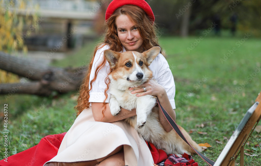 A girl with long red hair, in a red coat and beret gently hugs her corgi dog in an autumn park. Animal protection, dog walking, friendship, beautiful autumn picture