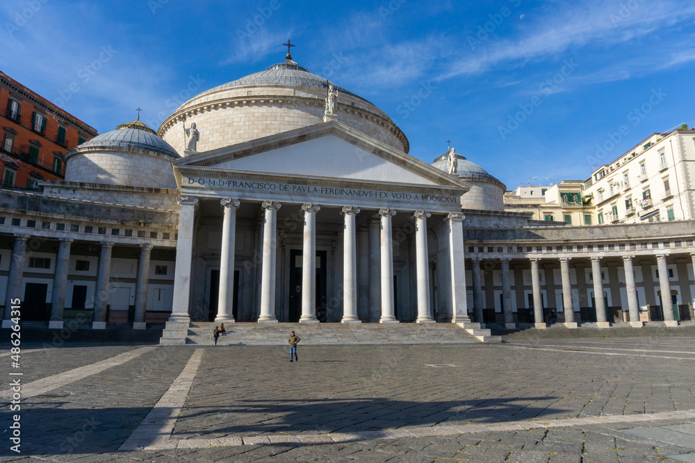 Piazza del Plebiscito in Naples, with the blue sky and some high clouds, with the colored houses behind. View of the basilica in the foreground.