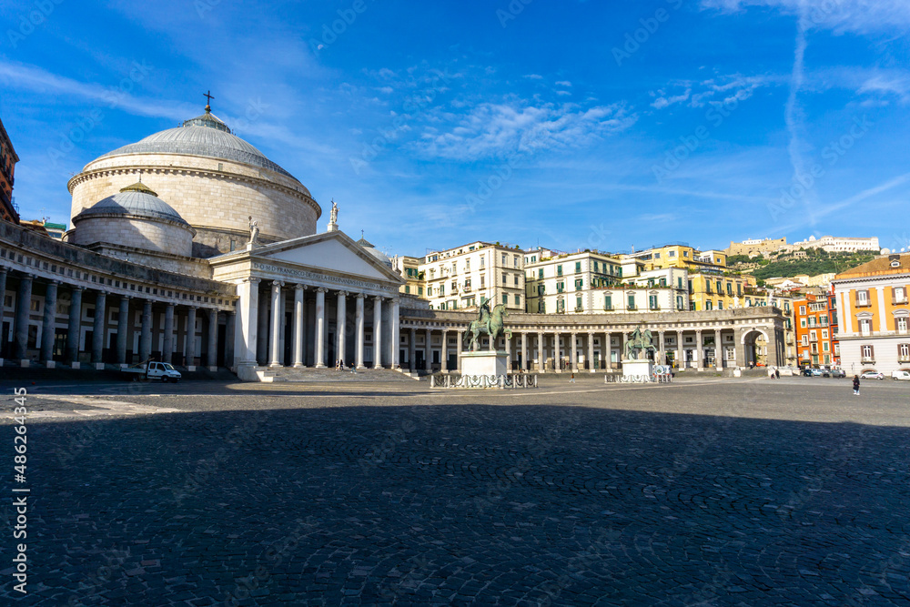 Piazza del Plebiscito in Naples, with the blue sky and some high clouds, with the colored houses behind and the Sant'Elmo castle. View of the basilica in the foreground.