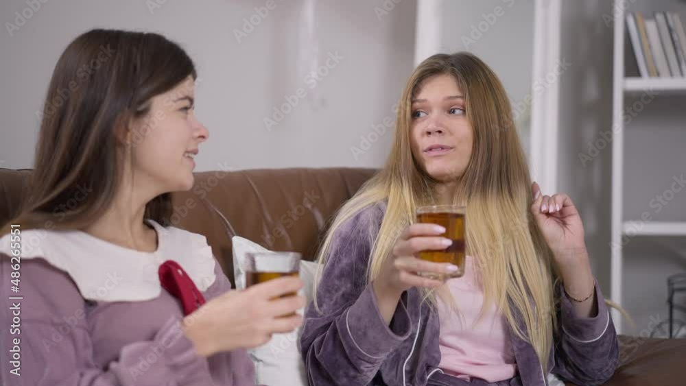 Two young women toasting drinking juice at pajama party smiling talking. Positive Caucasian female friends gossiping meeting indoors on weekend enjoying leisure. Lifestyle and happiness Two young women toasting drinking juice at pajama party smiling talking. Positive Caucasian female friends gossiping meeting indoors on weekend enjoying leisure. Lifestyle and happiness