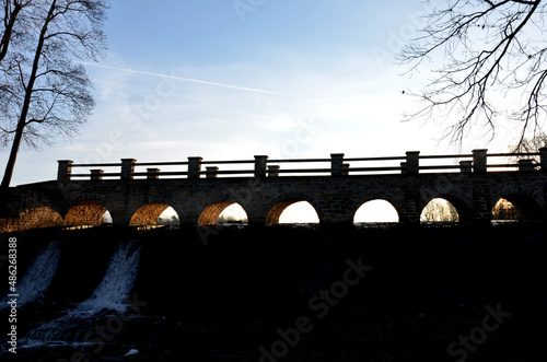 The dam of the breeding fish pond has a canal safety overflow similar to a weir. the water flows under a stone bridge with several arches. bridge on crown of dam with a wooden railing in silhoulete photo