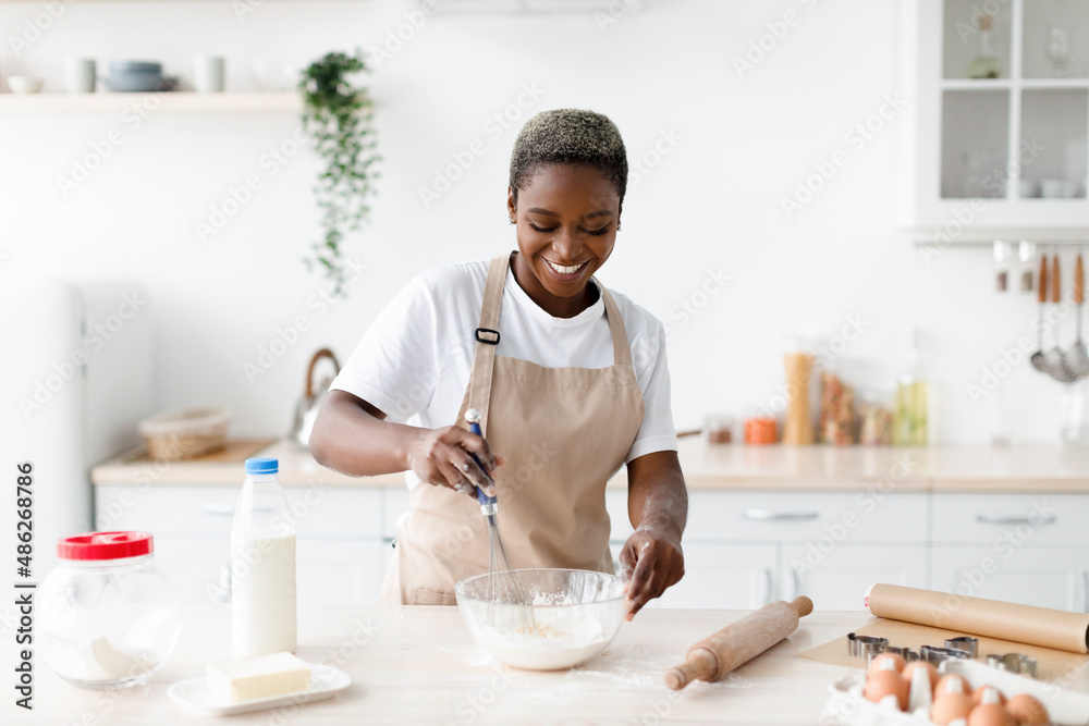 Glad happy young african american female in apron make dough for baking, prepares eat in scandinavian kitchen Glad happy young african american female in apron make dough for baking, prepares eat in scandinavian kitchen