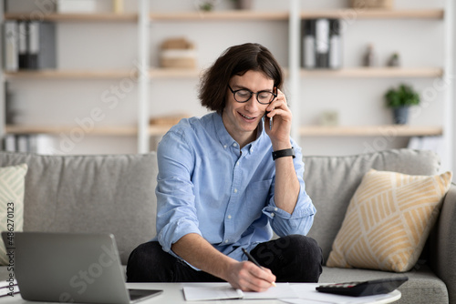 Successful young man talking on cellphone and taking notes, arranging job interview, sitting on sofa at home