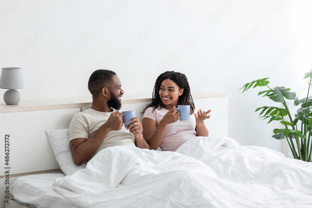 Cheerful young black husband and wife relaxing in bed with cups of coffee and talking, enjoy morning together Cheerful young black husband and wife relaxing in bed with cups of coffee and talking, enjoy morning together