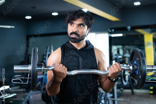 Young Indian bodybuilder busy doing biceps workout by lifting weight using barbells at gym - concept of musclebuilder exercising, healthy lifestyle and self care.