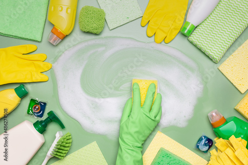 Home cleaning products. A hand in a green rubber glove wipes soap suds with a washcloth on a green background. Concept of cleanliness, cleaning services.