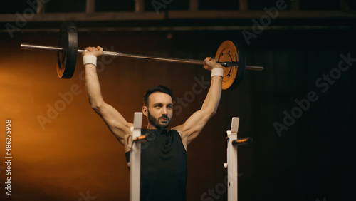Muscular sportsman training with barbell near stand on dark background