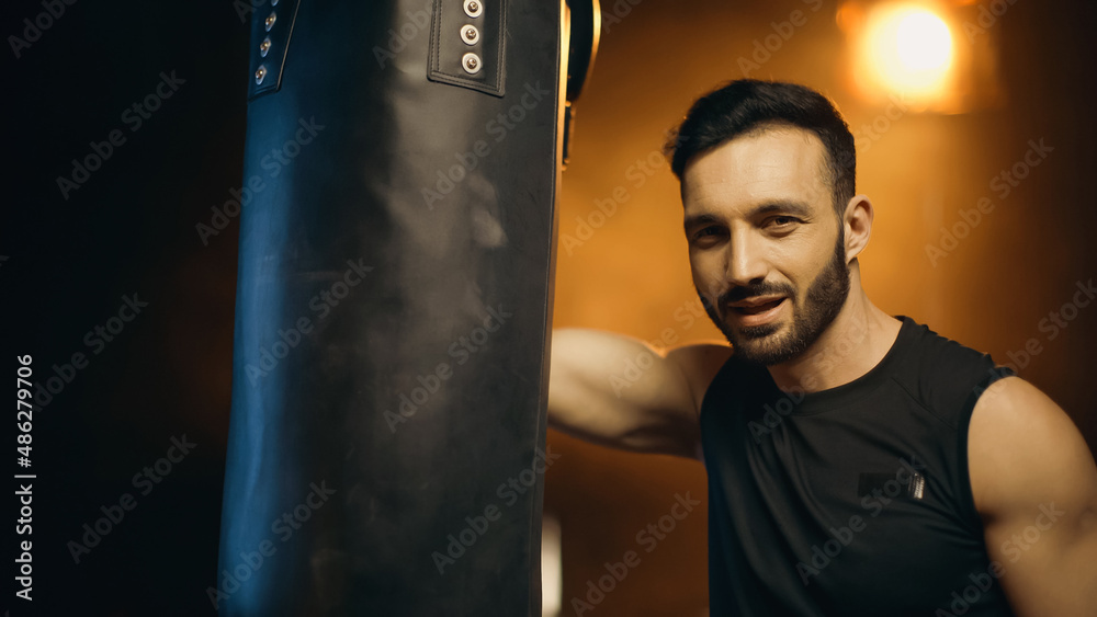 Smiling sportsman looking at camera near punch bag on dark background