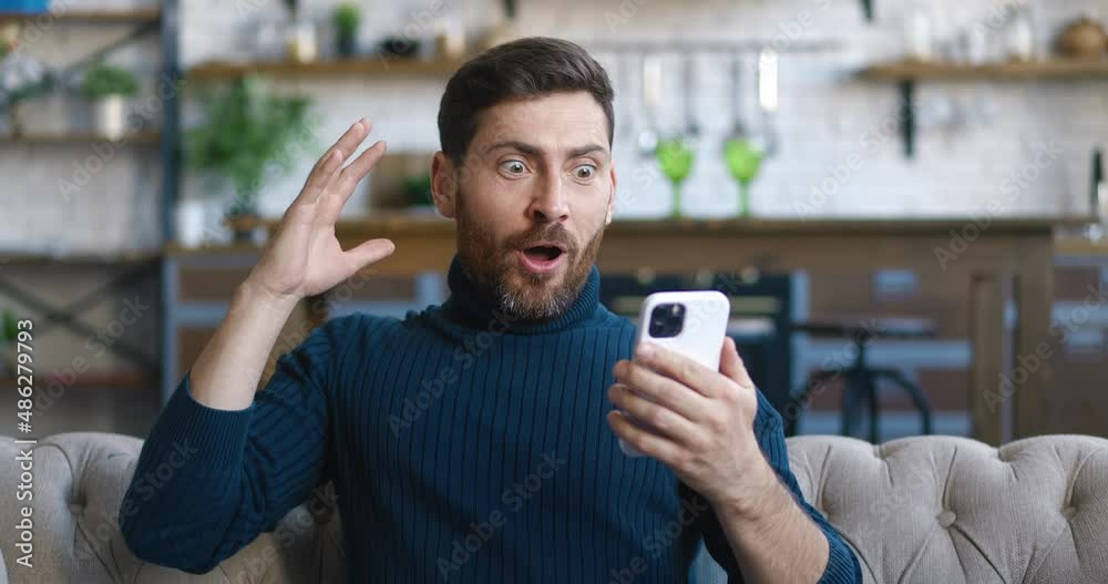 Excited and pleasantly surprised handsome man rejoices at the message on his smartphone. Young male looking at mobile phone and feel amazed happy and saying WOW on home background. Excited and pleasantly surprised handsome man rejoices at the message on his smartphone. Young male looking at mobile phone and feel amazed happy and saying WOW on home background.