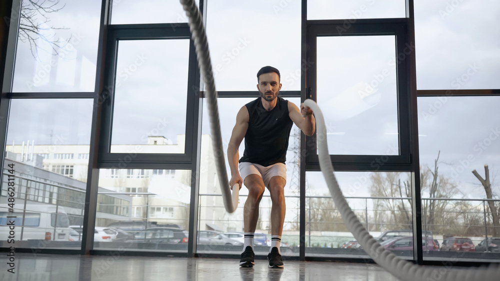 Obraz premium Athletic sportsman training with blurred battle rope in gym