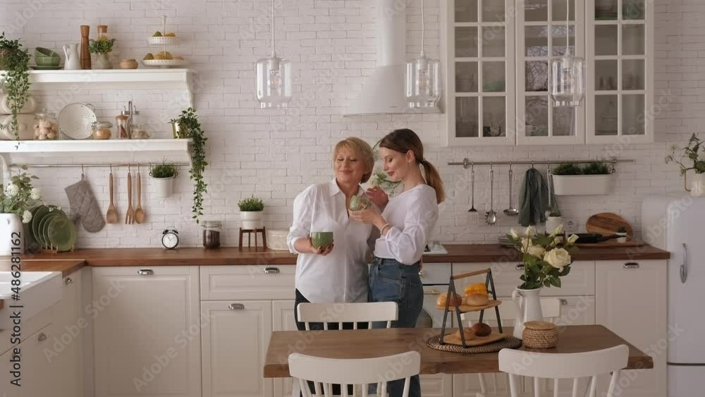 Portrait of a happy elderly mother with her beloved daughter at home in the kitchen, they are dressed in comfortable jeans and shirts. An old mother embraces her adult daughter. Portrait of a happy elderly mother with her beloved daughter at home in the kitchen, they are dressed in comfortable jeans and shirts. An old mother embraces her adult daughter.