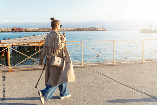 Young adult trendy stylish beautiful caucasian happy smiling woman enjoy walking by Yalta sea embankment on warm sunny day. Female person portrait wear jeans biege trench coat on urban city street photo
