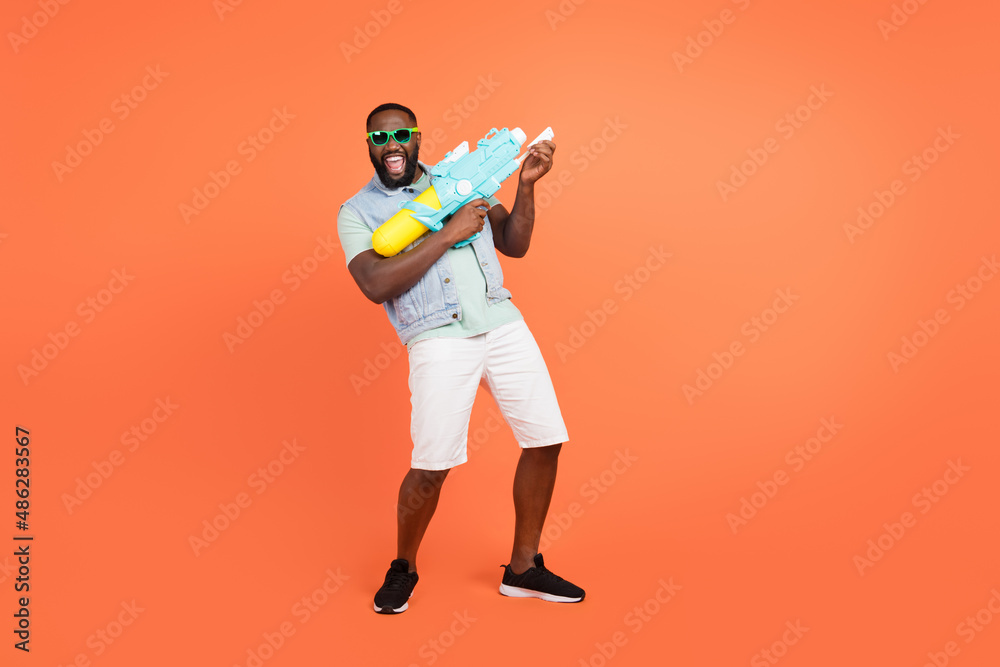 Photo of excited guy wear denim vest dark eyewear playing water gun empty space isolated orange color background Photo of excited guy wear denim vest dark eyewear playing water gun empty space isolated orange color background
