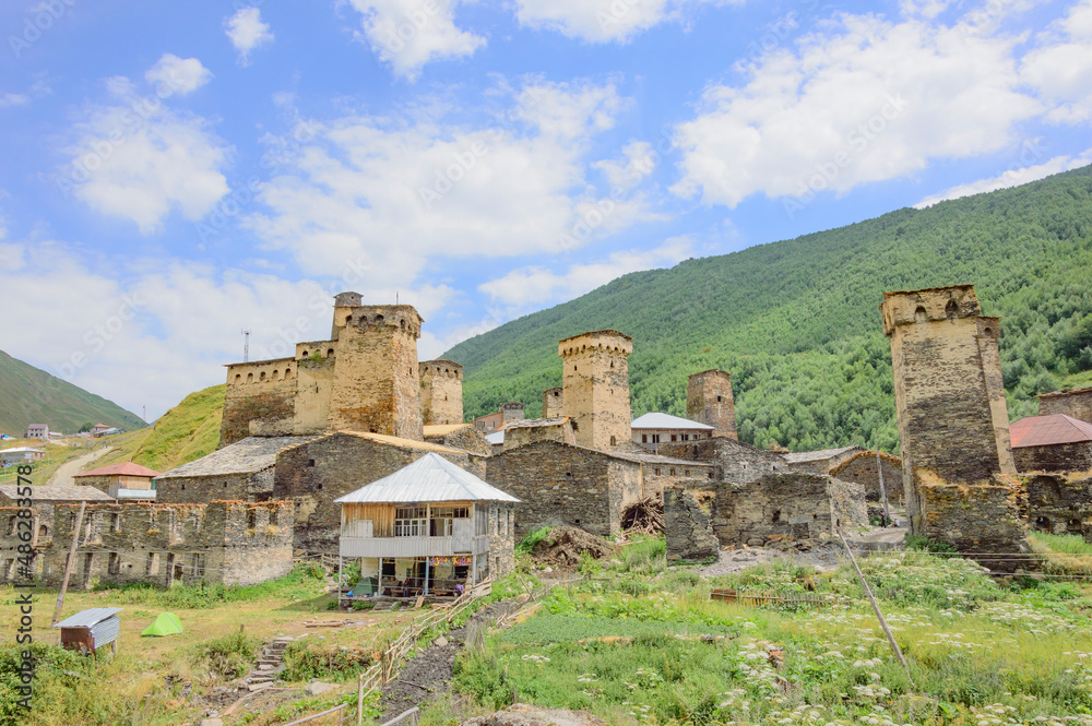 View of Ushguli fortified village in Svanetia, Georgia. Grey, brown and biege medieval stone towers and old houses, green and yellow dry grass, green mountains and blue sky with clouds