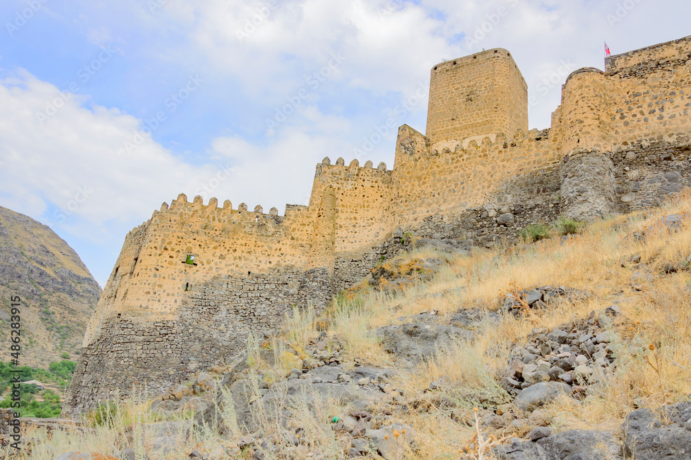 Khertvisi fortress in Georgia, Caucasus. Ancient weathered medieval yellow stone walls and towers, grey cliff, blue sky with clouds, dry yellow grass
