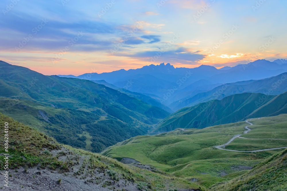 View of the summer sunset above the caucasian mountains from the Bear Cross mountain pass in Khevsureti, Georgia. Blue and orange sky with clouds, green and blue haze maountains, road to Shatili