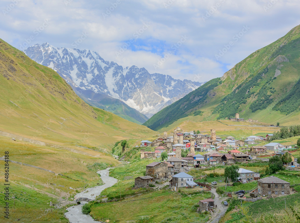 Hilltop view of Ushguli medieval fortified village in Svaneti, Georgia. Old stone towers and houses, green and yellow dry grass, road, green and rocky snow mountains and blue sky with clouds