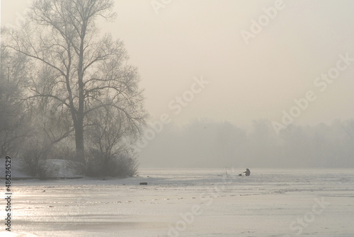 A blurry winter landscape with a frozen river, trees in hoarfrost and unrecognizable man fishing on the ice.