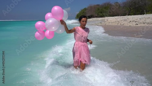 Cute happy woman in pink with balloons walking in the splashing ocean water, birthday party on the seaside 