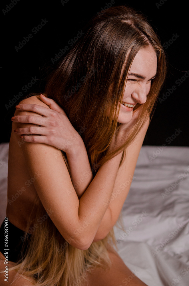 Beautiful young woman with blonde hair in black underwear is sitting on bed in aesthetic poses, on a black background Beautiful young woman with blonde hair in black underwear is sitting on bed in aesthetic poses, on a black background