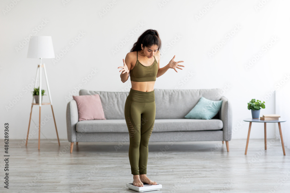 Fototapeta premium Emotional Indian woman standing on scales, feeling disappointed about weight loss, failing to achieve slimming result