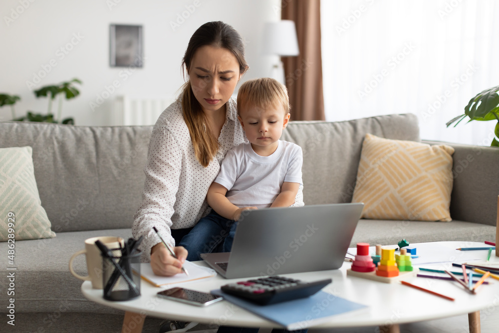 Mom working remotely on laptop computer while taking care of baby, mother on maternity leave trying to freelance Mom working remotely on laptop computer while taking care of baby, mother on maternity leave trying to freelance