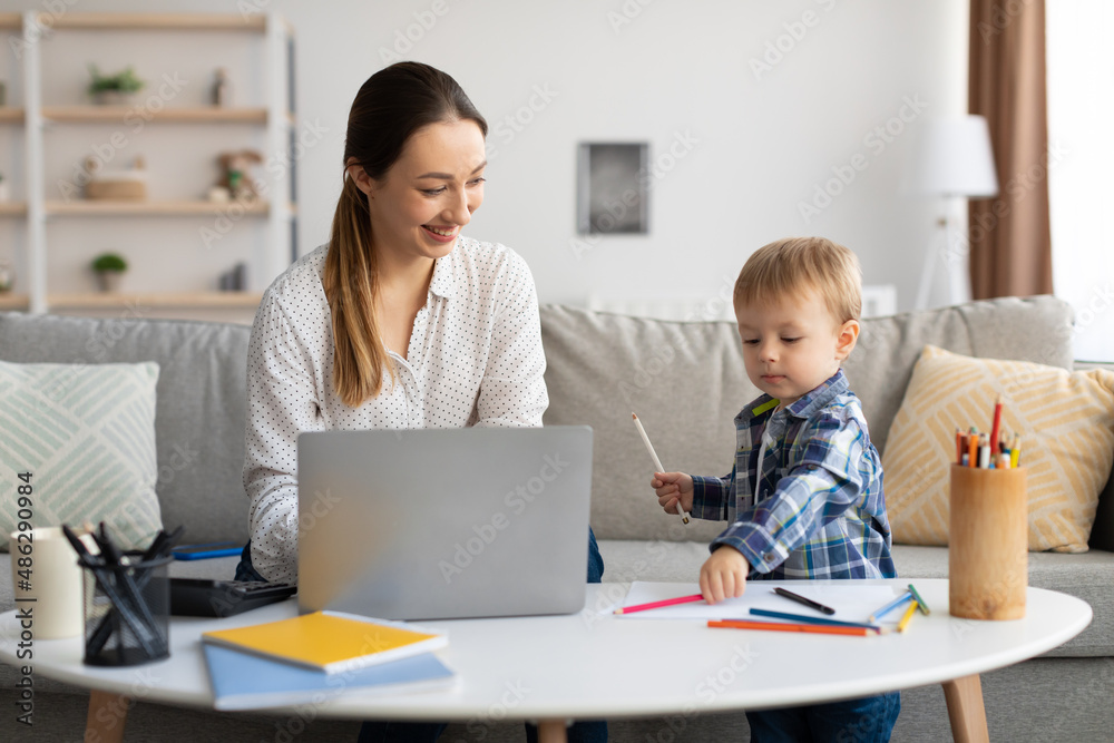 Work at maternity leave. Happy young mom working on laptop and looking at her toddler son drawing with pencils Work at maternity leave. Happy young mom working on laptop and looking at her toddler son drawing with pencils