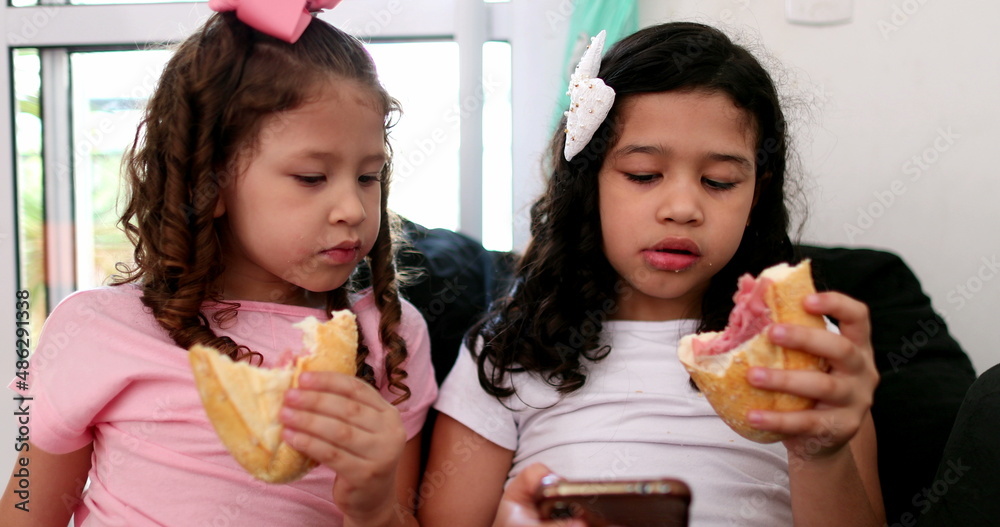 Two little girls hypnotized by cellphone while eating sandwich snack Two little girls hypnotized by cellphone while eating sandwich snack