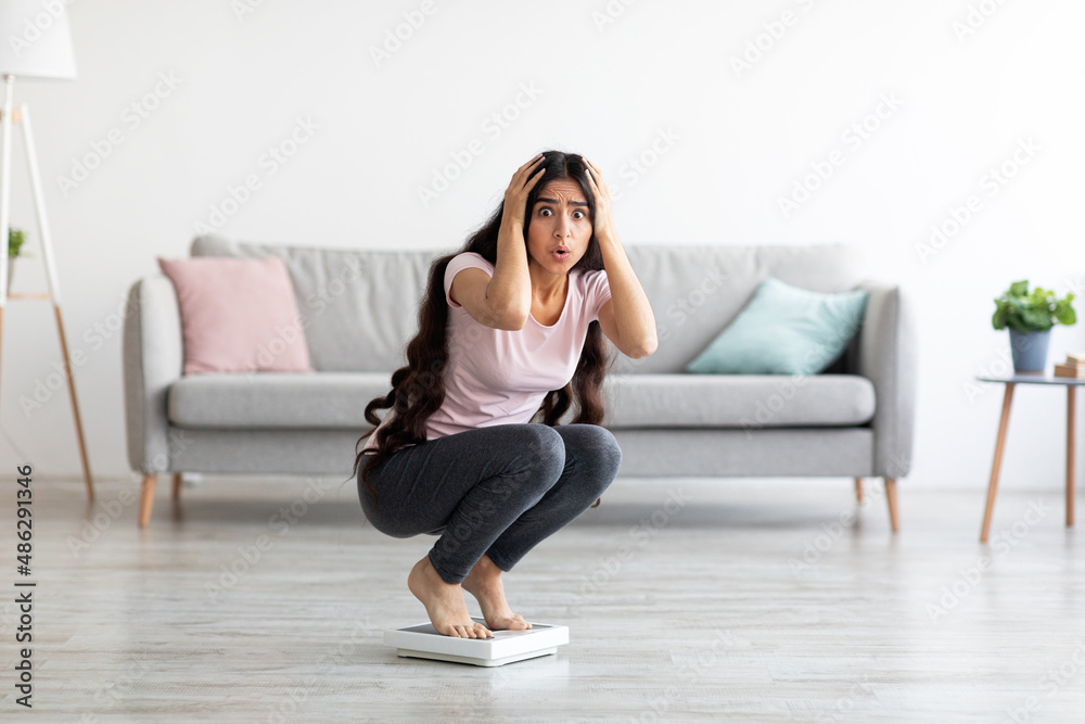 Shocked young Indian lady sitting on scales, terrified of weight gain at home, free space. Healthy living concept