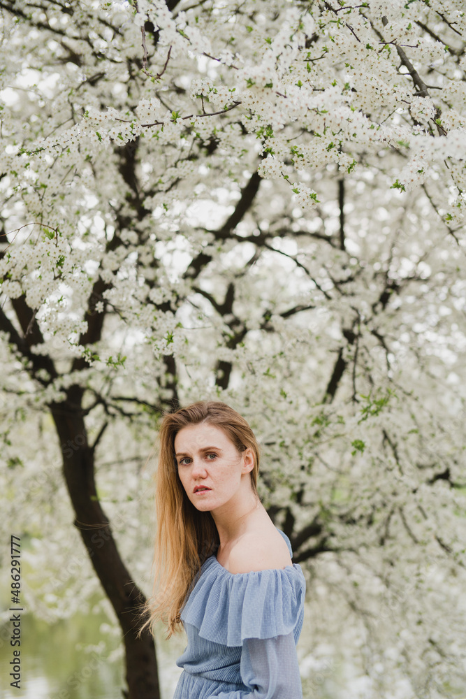 Young pretty caucasian blonde woman with freckles wearing natural makeup in light blue dress near the beautiful blooming spring tree by the lake. Youth, freshness, beauty, happiness, emotions concept. Young pretty caucasian blonde woman with freckles wearing natural makeup in light blue dress near the beautiful blooming spring tree by the lake. Youth, freshness, beauty, happiness, emotions concept.