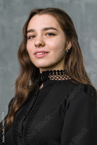 Studio portrait of young beautiful girl with dark hair and brown eyes. Grey wall in background. Black dress.