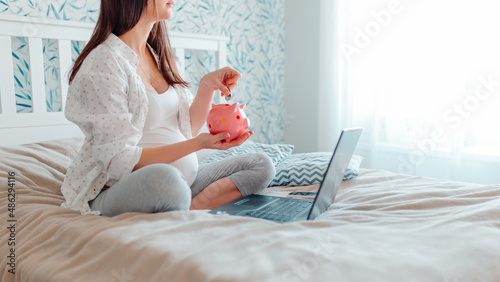Pragnant woman puts coins into piggy bank. Pregnant with laptop on bed. Pregnant woman online shopping.