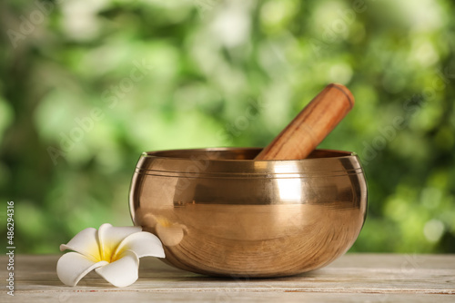 Golden singing bowl, mallet and flower on white wooden table outdoors