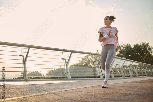 Young athletic woman, jogger, runner practicing sport outdoors, running fast along the bridge over the sunrise sky and river background. Healthy lifestyle concept. Workout jogging activity