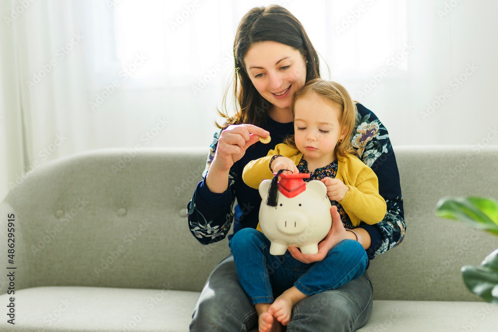 Cheerful Mother Teaching Little baby How To Save Money Holding Piggybank Sitting On Sofa At Home Cheerful Mother Teaching Little baby How To Save Money Holding Piggybank Sitting On Sofa At Home