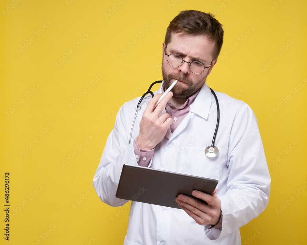 Pensive doctor with a stethoscope around neck holds a tablet in hands and a stylus in his lips. Pensive doctor with a stethoscope around neck holds a tablet in hands and a stylus in his lips.