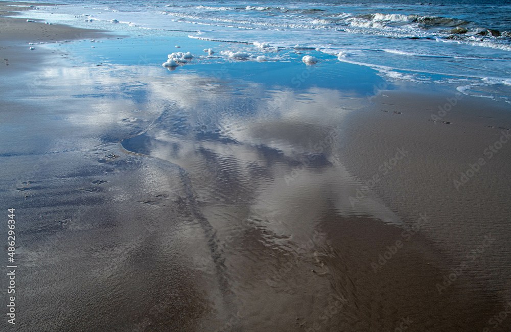 Weißer Schaum am Strand und Wolken spiegeln sich im Wasser Stock Photo