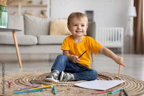 Portrait of happy toddler boy sitting on carpet with colorful pencils and sheet of paper, drawing at home, copy space
