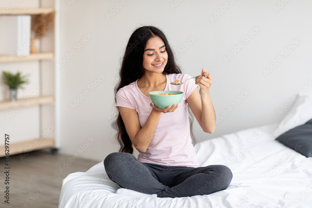 Full length portrait of beautiful millennial Indian lady having cereal with milk while sitting on bed at home Full length portrait of beautiful millennial Indian lady having cereal with milk while sitting on bed at home
