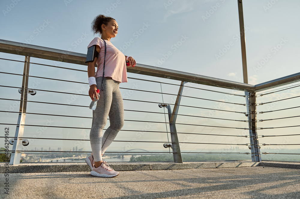 Full length portrait of a sportswoman, female athlete with a skipping rope, standing on the modern glass city bridge, relaxing after cardio workout outdoor. Sport, fitness, active lifestyle concept