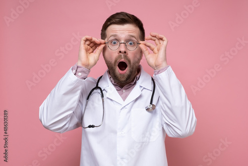 Young doctor in a white coat, with a stethoscope around neck, opened mouth in shock and looks with big eyes holding glasses. 