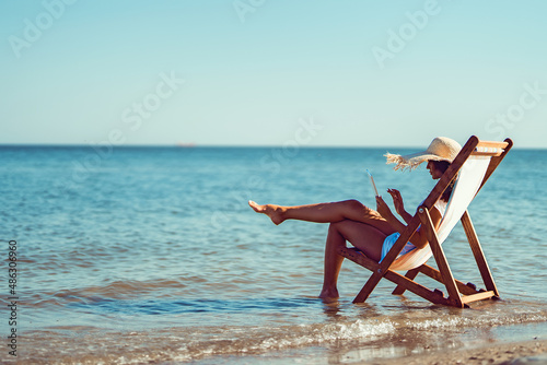 Back view girl in sunhat working at laptop while lying on the beach chaise longue at the seaside