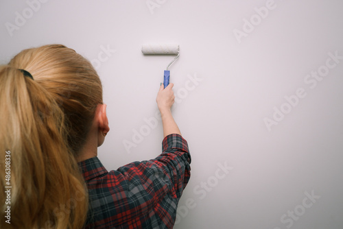 Painting works. A woman in a plaid shirt paints a light wall with a paint roller. View from the back to the hand with the tool.