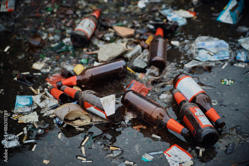 A large pile of household garbage on the sidewalk. Used glass bottles and paper packaging are lying on the ground