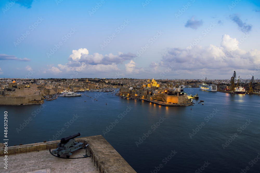Fototapeta premium Malta panorama of fortified Three Cities with moored ships and clouds above. Evening view of Vittoriosa, Senglea and Cospicua across the Grand Harbour, seen from Upper Barrakka Gardens in Valletta.