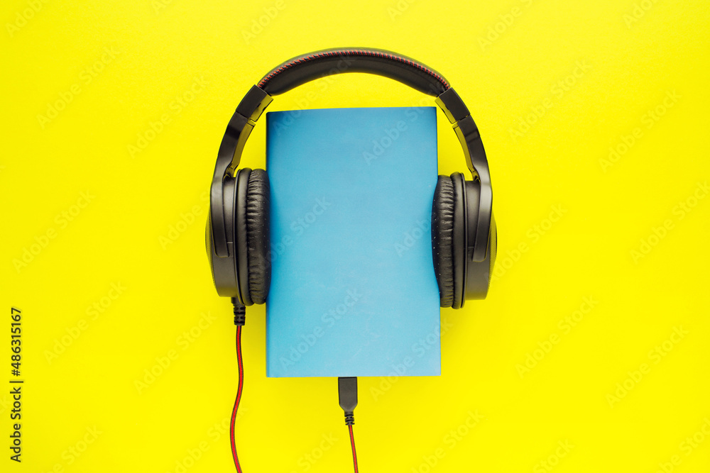 Book in a blue cover and black headphones on a bright yellow background, top view, close-up. Conceptual image of listening to an audiobook, reading, education Book in a blue cover and black headphones on a bright yellow background, top view, close-up. Conceptual image of listening to an audiobook, reading, education