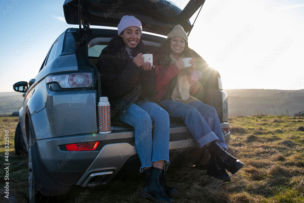 Portrait of female friends sitting in car trunk and drinking tea