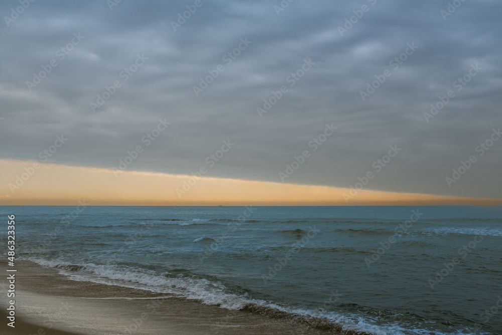 Sunlight tries to break through the thick blanket of clouds creating suggestive zig zag shapes between the shore, the water and the sky. Mediterranean Sea on a cloudy day
