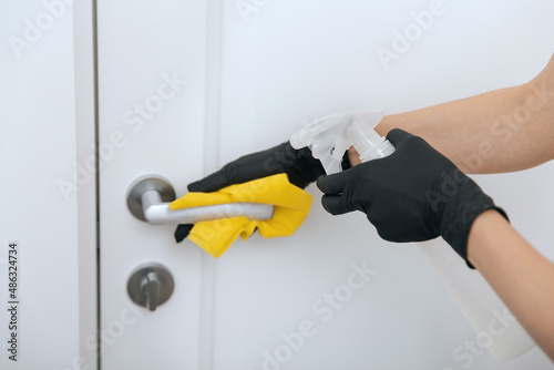 Cleaning door handle with yellow wipe in black gloves and sanitizer spray. Disinfection in hospital and public spaces against corona virus. Woman hand using towel for cleaning.